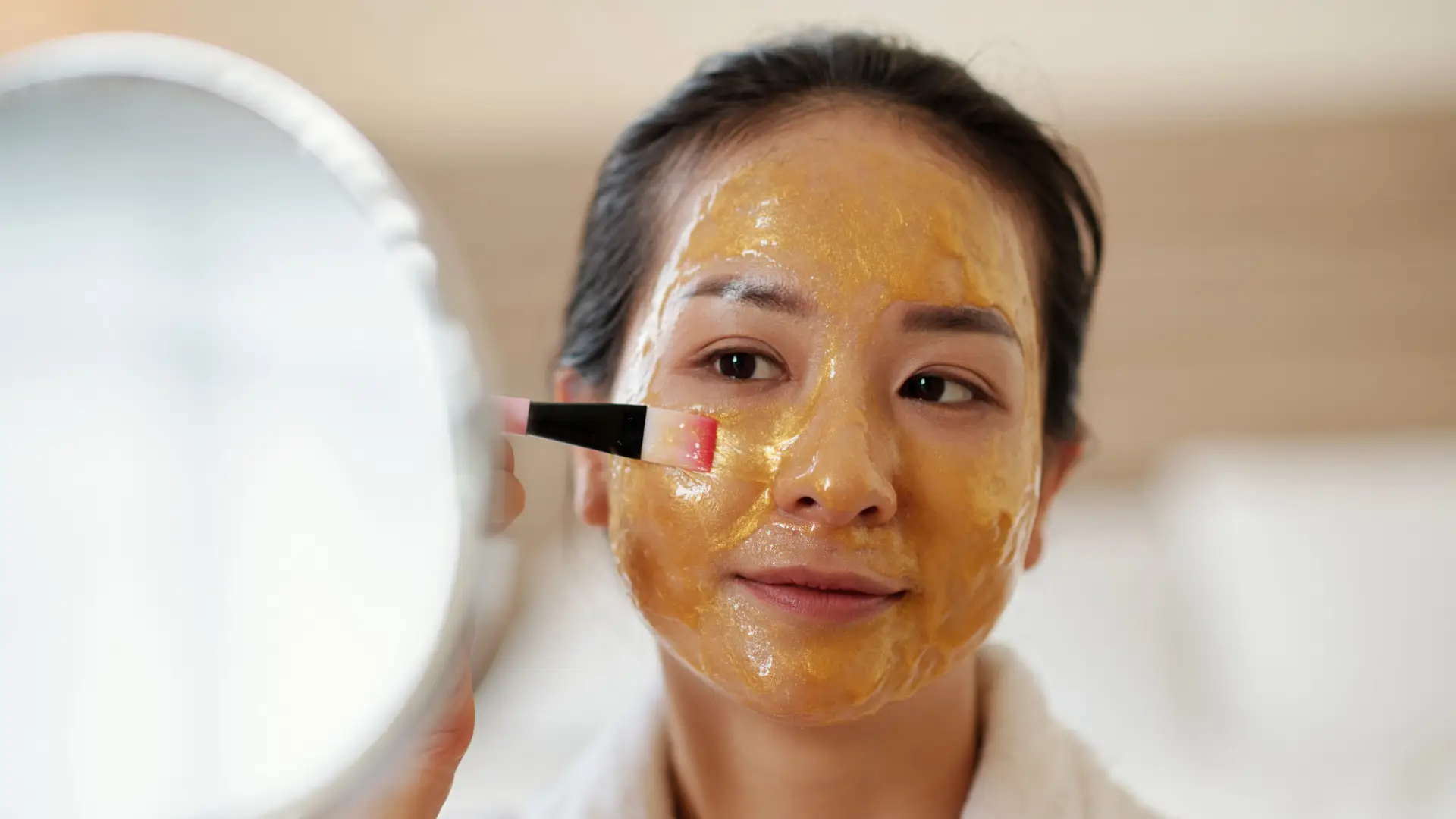 Femme appliquant un masque de beauté doré au miel sur son visage à l'aide d'un pinceau devant un miroir.